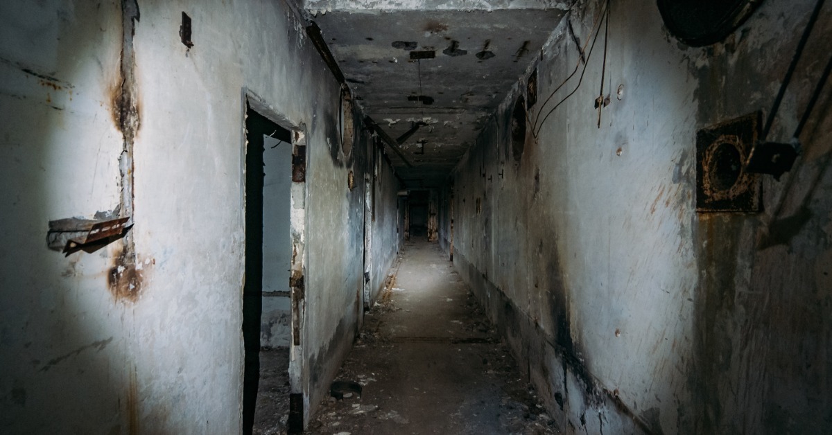 A very dark and creepy looking corridor were the walls are covered in rust and grime. There are doorways down the hall.