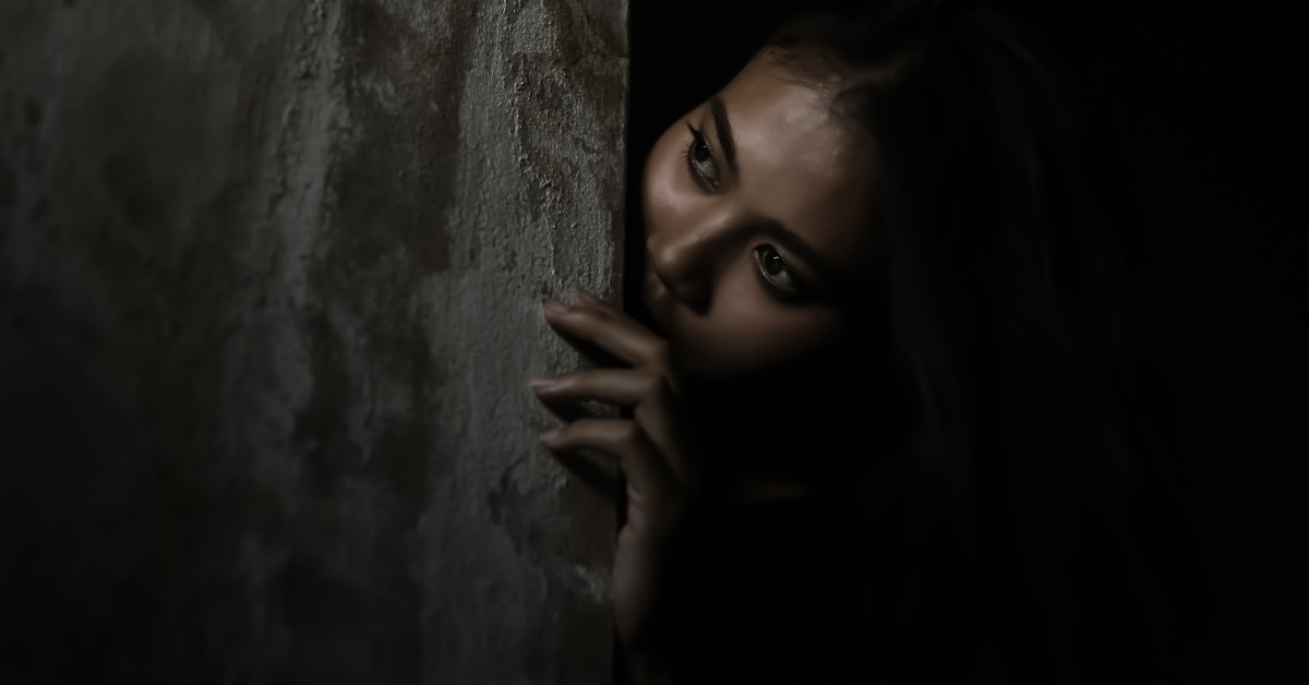 The head of a scared looking woman peeking around the corner of a wall in a very dimly lit corridor.